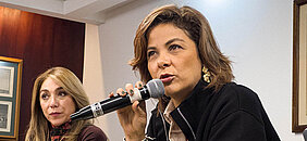 En la imagen se puede ver a la ministra Jhenifer Mojica, ella se encuentra hablando por medio de un micrófono en una sala, atrás de ella se encuentra otra mujer mirando fijamente al receptor.