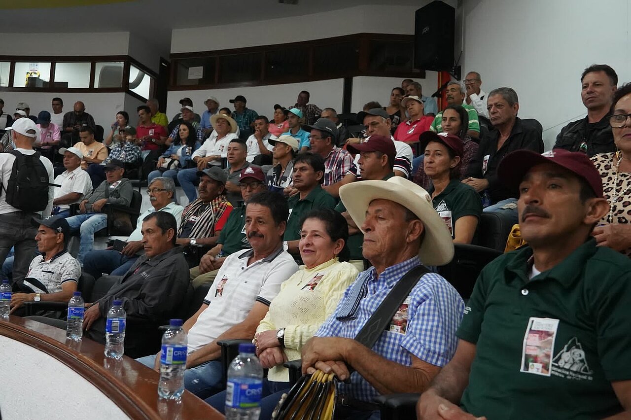 La imagen representa a un grupo numeroso de personas reunidas. Muchos de los espectadores llevan sombreros de vaquero, lo que indica un posible tema cultural o regional. Los espectadores están sentados en sillas, dispuestas en filas, y hay varias botellas colocadas alrededor, probablemente con bebidas para los asistentes.