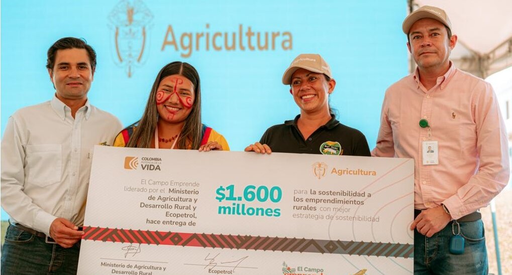 La imagen muestra a un grupo de cuatro personas de pie, posando para una foto. Todos sonríen y parecen felices. Dos de las personas sostienen un gran cheque. El grupo está formado por dos hombres y dos mujeres, todos vestidos con ropa informal. Los dos hombres sostienen el cheque, mientras que las mujeres permanecen de pie a su lado, lo que contribuye al ambiente festivo.