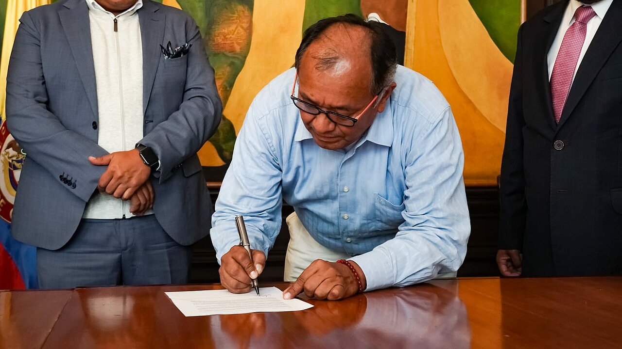 En la imagen se muestra a un señor firmando un documento, al lado de el se encuentran dos señores de traje, de fondo un cuadro con colores amarillo y verde, y de fondo se puede ver la bandera Colombiana