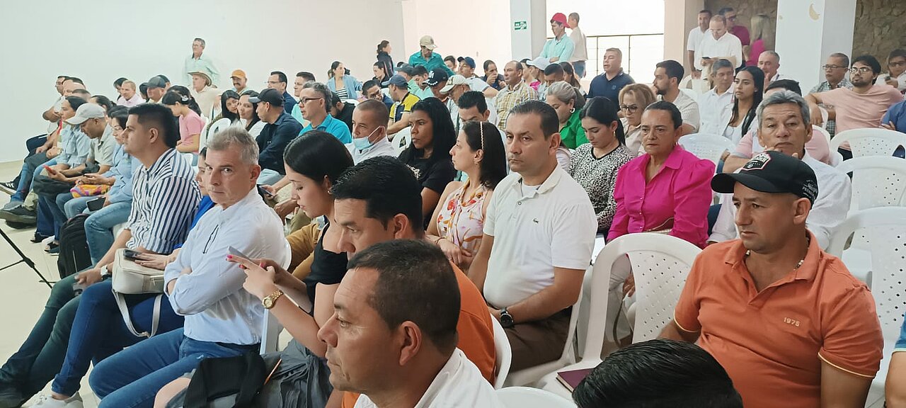 La imagen muestra a un grupo de personas sentadas en filas, mirando hacia la parte delantera de la sala, donde podría estar teniendo lugar un orador o una presentación. Los asistentes parecen estar participando en una reunión o conferencia, como sugieren sus posturas atentas y el entorno.