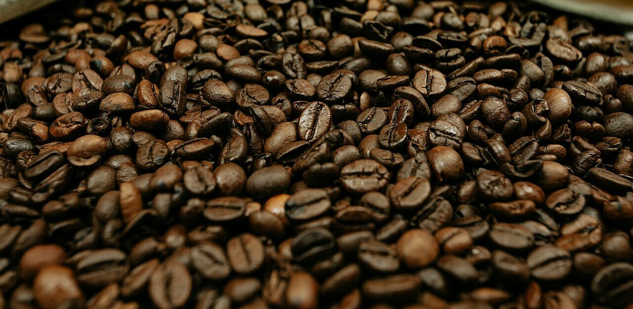 La imagen muestra un gran montón de granos de café, algunos verdes y otros tostados. Los granos están esparcidos en varias direcciones, creando un conjunto visualmente atractivo y abundante. El montón está metido en una bolsa blanca, lo que acentúa aún más el contraste entre los granos y el fondo. El primer plano de los granos resalta sus variaciones de textura y color, mostrando la diversidad y riqueza de la selección de café.