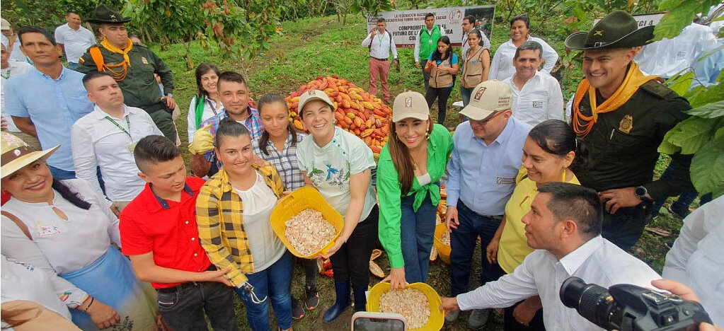 Esta imagen muestra a un grupo de personas reunidas al aire libre, posiblemente en una granja o evento agrícola. Sostienen bandejas amarillas con lo que parece ser un tipo de grano o semillas. Las personas sonríen y parecen posar para la foto, lo que indica un ambiente positivo y amistoso.