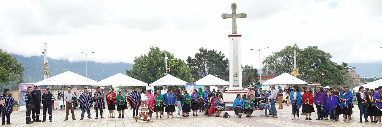 Esta imagen muestra a un grupo de personas reunidas en una plaza pública.  Las personas están vestidas con diversos atuendos, algunos de los cuales parecen ser tradicionales o culturales, lo que indica que podría tratarse de un acontecimiento o celebración especial. El cielo está nublado y el ambiente general parece el de una reunión comunitaria o una ceremonia.