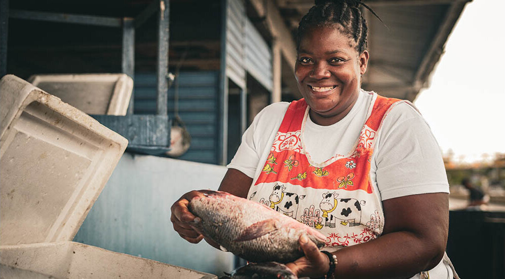 En la imagen se ve a una persona sosteniendo un gran pez. La persona parece sonreír y parece estar en un entorno que podría ser un mercado o un lugar donde se vende o se prepara pescado. El pescado es bastante grande y la persona lo sostiene para mostrarlo.