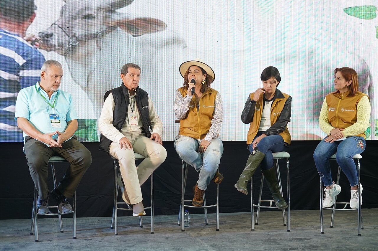 La imagen muestra a un grupo de cinco personas sentadas en taburetes frente a una gran pantalla. Parece que están participando en un debate o una presentación, y una de ellas habla por un micrófono. El grupo está formado por tres hombres y dos mujeres, todos vestidos con ropa informal.