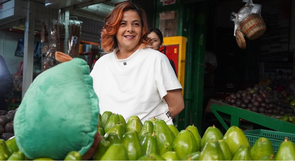 En la imagen aparece la Ministra de pie detrás de un expositor de frutas verdes, que parecen ser aguacates. Sonríe y parece estar en un mercado o tienda, posiblemente vendiendo los productos.