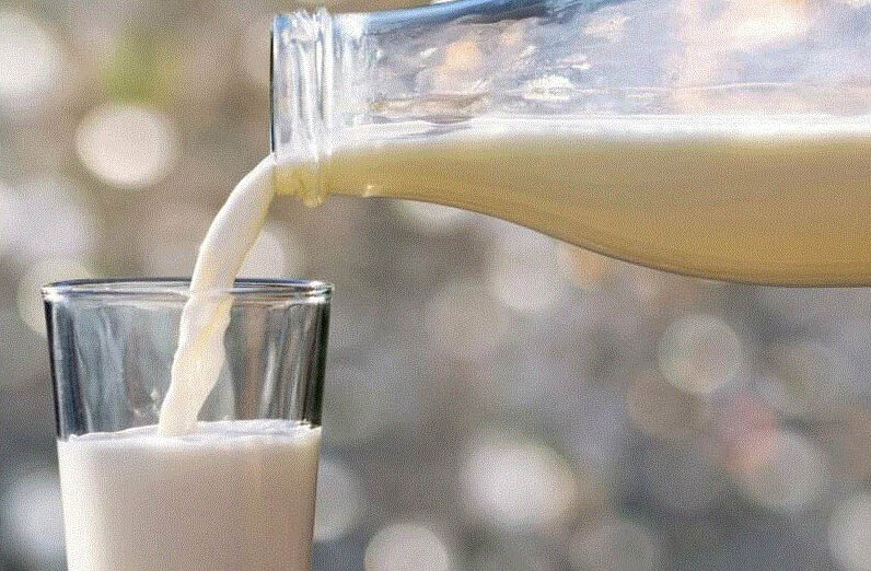 La imagen muestra un vaso de leche que se llena con una botella. La leche fluye de la botella al vaso, creando un chorro suave y blanco. El fondo está difuminado, lo que sugiere una escasa profundidad de campo, centrando la atención del espectador en la leche y en la acción de verterla. La iluminación parece suave y difusa, lo que podría indicar un interior con luz ambiental.