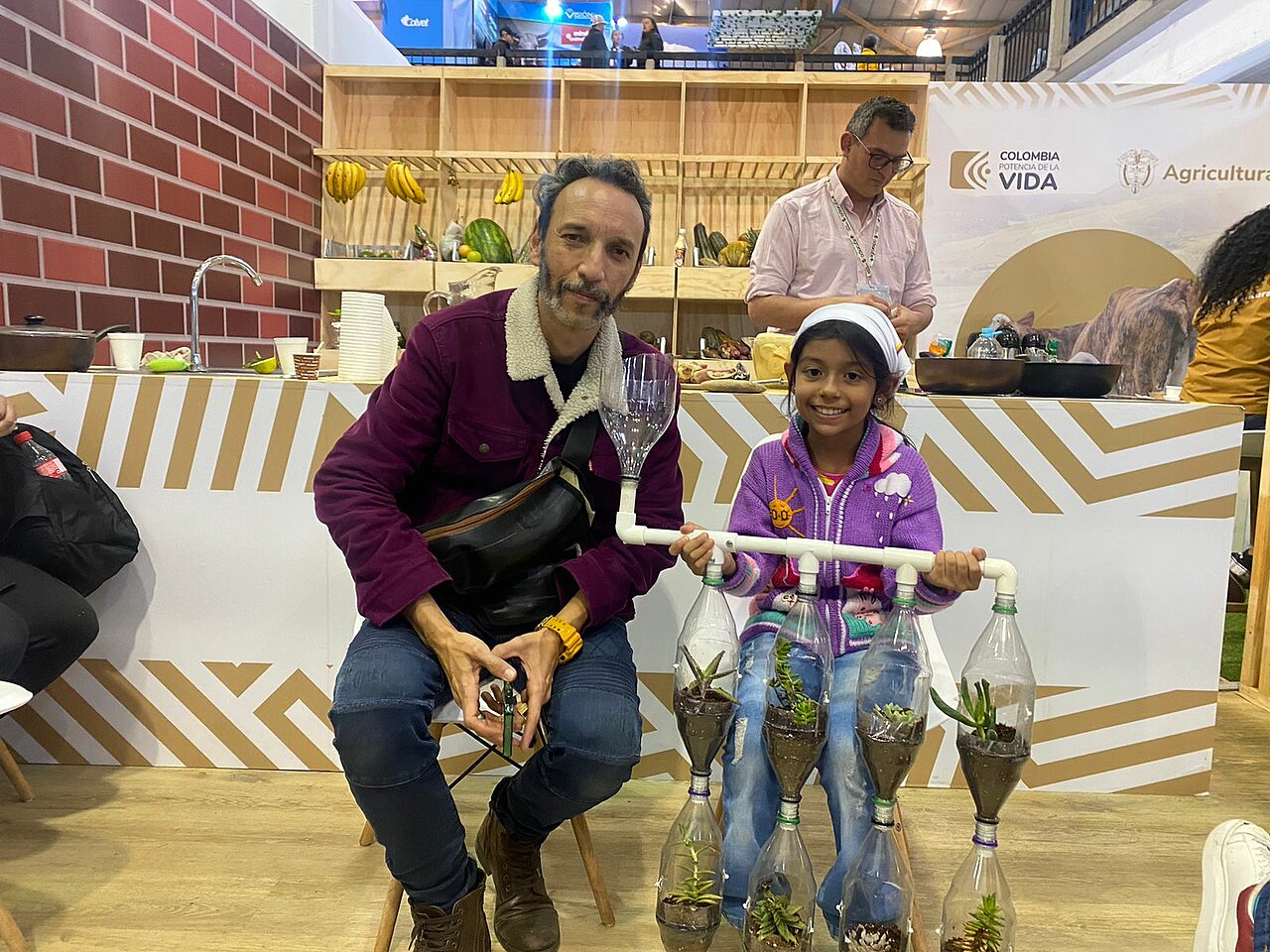 La imagen muestra a un hombre y una niña sentados juntos en un banco, rodeados de varios objetos. El hombre sostiene una copa de vino y la niña una jeringuilla de juguete. Hay varias botellas y vasos esparcidos por la escena, así como un bolso colocado cerca.  Al fondo, hay algunas personas más, posiblemente realizando diferentes actividades. La escena parece ser una reunión informal o un acto social en el que la gente disfruta de la compañía de los demás.