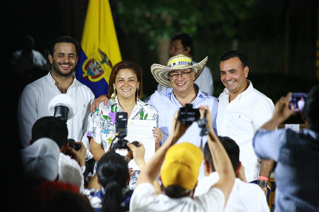 En la imagen, hay cuatro individuos de pie frente a una multitud de personas que están haciendo fotos y grabando vídeos. La persona del centro sostiene un documento, lo que sugiere que podría estar recibiendo un premio o un reconocimiento.