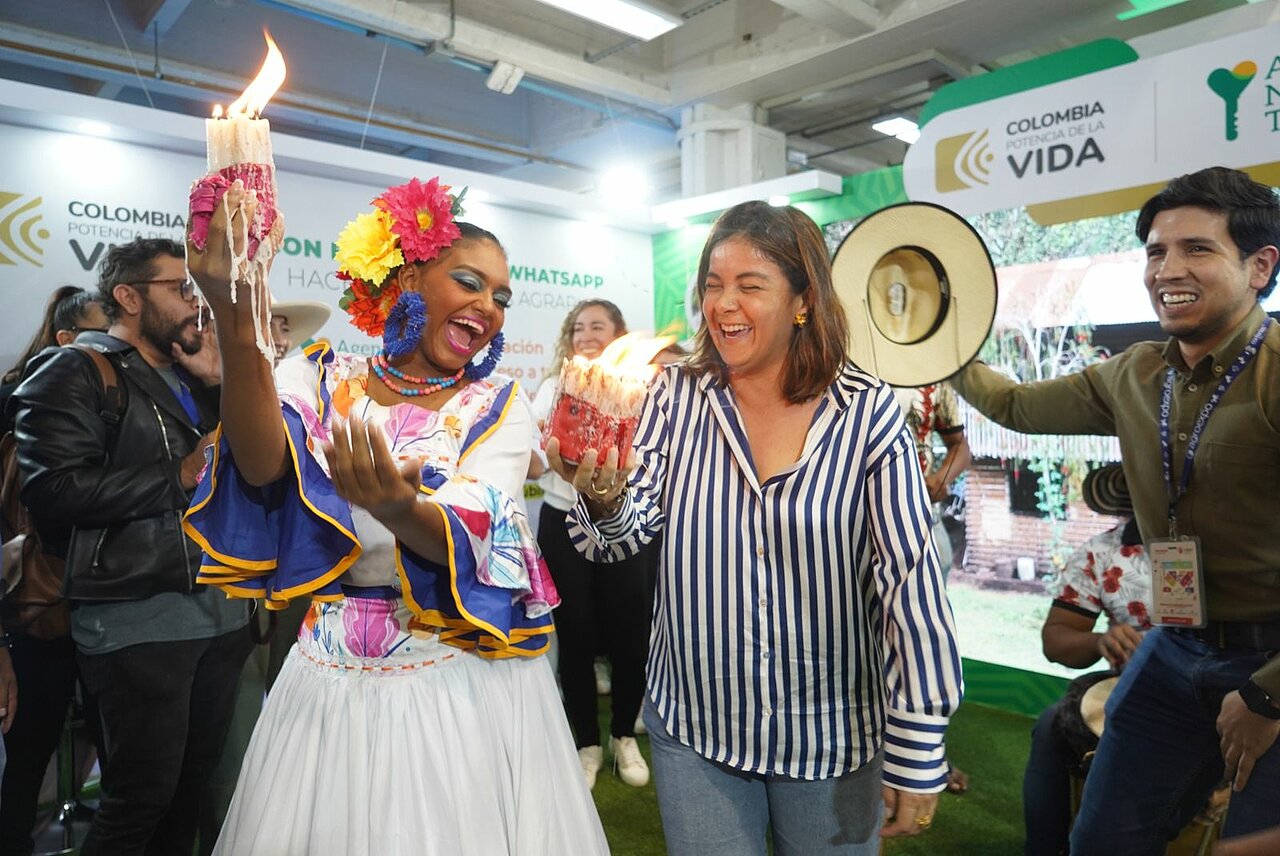 La imagen representa una animada escena con una mujer vestida con la ropa tradicional colombiana, sosteniendo una vela encendida en cada mano. La rodea un grupo de personas, entre ellas La Ministra Jhenifer Mojica, vestida con camisa de rayas, que sonríen y disfruta del ambiente festivo. La mujer vestida con el traje tradicional parece ser el centro de atención, ya que sostiene las velas y se relaciona con la multitud.