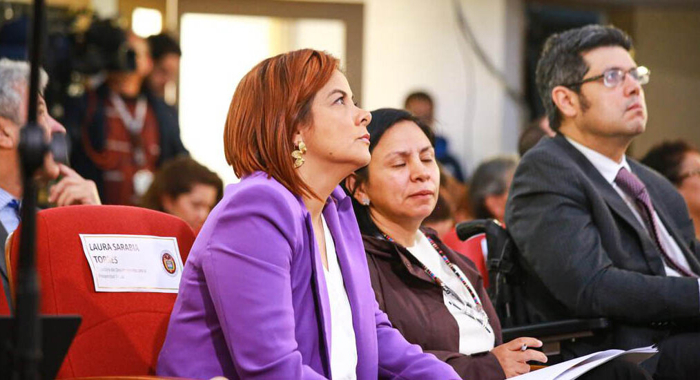 En la imagen se ve a tres personas sentadas en lo que parece ser un entorno formal o profesional. La persona en primer plano lleva una americana morada y mira hacia un lado con expresión pensativa. La persona del centro lleva una blusa blanca y tiene una expresión seria o contemplativa. La persona del fondo lleva un traje oscuro y también mira a un lado, posiblemente a algo o alguien que no se ve en la foto.  Traducción realizada con la versión gratuita del traductor DeepL.com