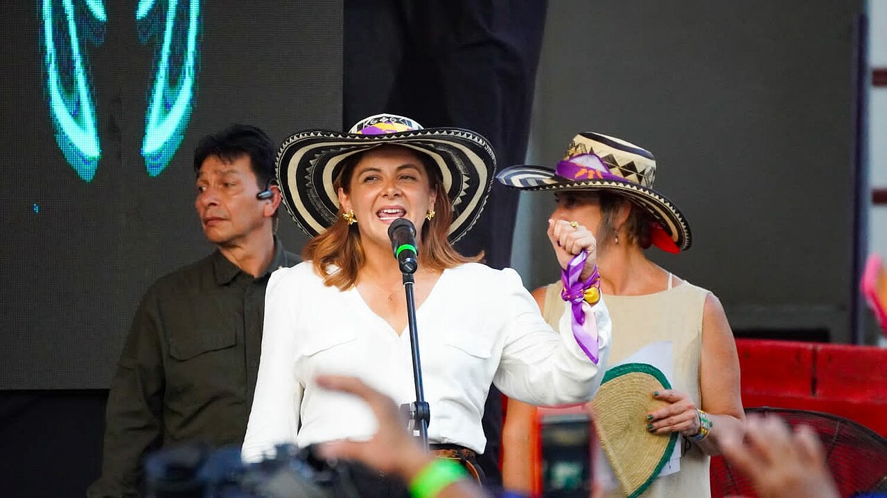 La imagen muestra a una mujer de pie en un escenario, con camisa blanca y sombrero. Sostiene un micrófono y parece estar hablando, por él. Hay otras dos mujeres en la escena, una a la izquierda de la mujer del micrófono y otra a la derecha. Ambas llevan sombrero.  Al fondo hay dos hombres, uno detrás de la mujer del micrófono y otro detrás de la mujer de la derecha. 