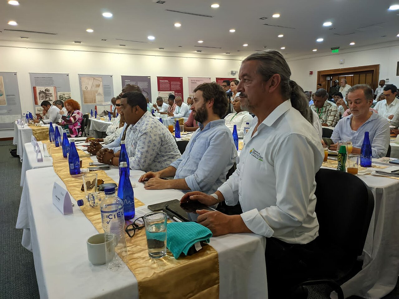En la imagen se ve a un grupo de personas sentadas a una larga mesa en lo que parece ser una conferencia o reunión. Las personas están vestidas con ropa informal de negocios, lo que sugiere un entorno profesional. Están mirando hacia la parte delantera de la sala, donde podría haber un orador o una presentación.