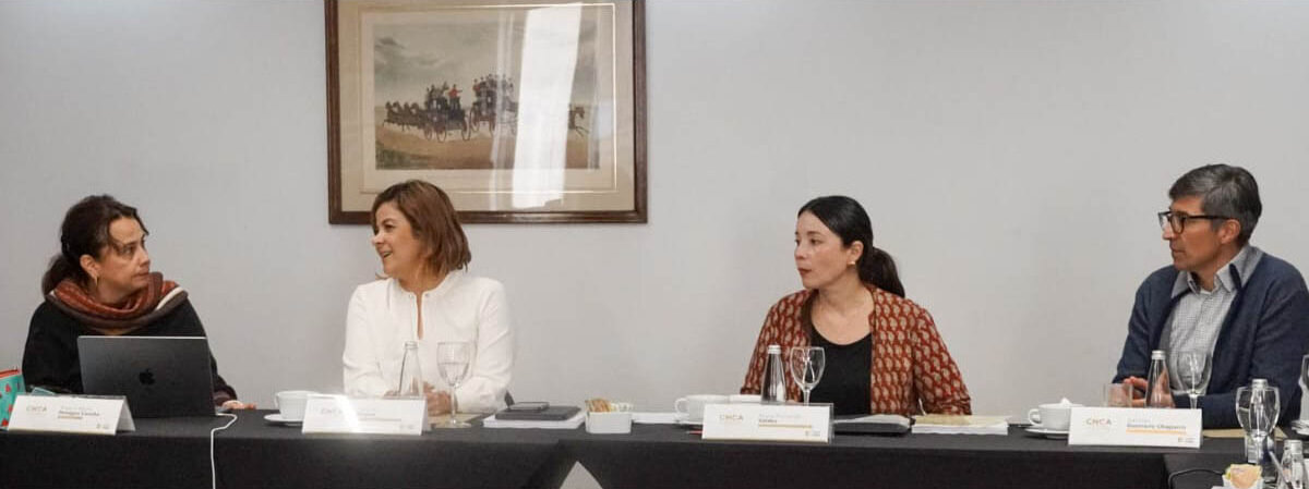 En la imagen, hay cuatro personas sentadas a una mesa, que parece ser un panel o una reunión. Todos visten traje, lo que sugiere un ambiente formal o profesional. También hay portátiles, micrófonos y otros objetos sobre la mesa, lo que refuerza la idea de que se trata de un acto o reunión formal.