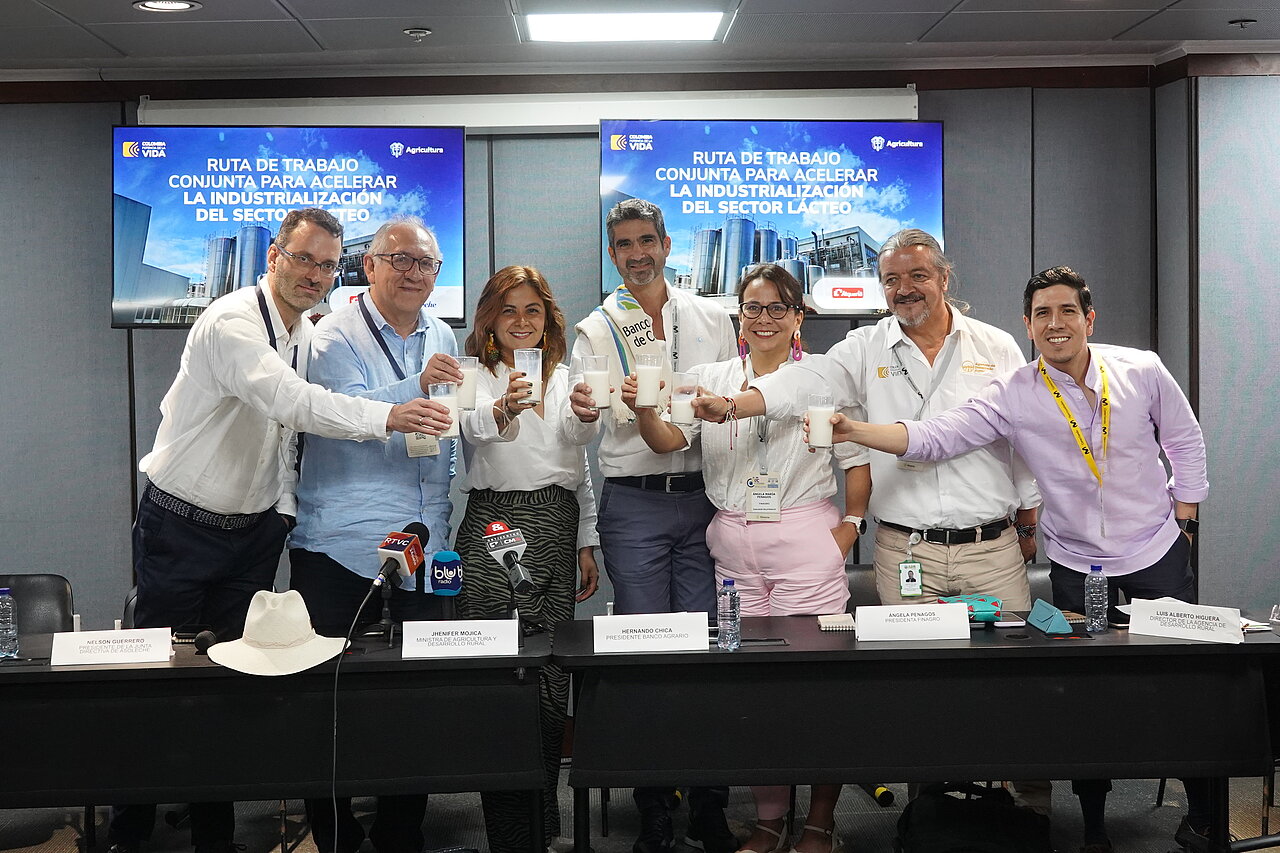 La imagen muestra a un grupo de personas reunidas en una sala de conferencias, posando para una foto. Hay nueve personas en total, de pie alrededor de una larga mesa. Todos sostienen sus teléfonos móviles, posiblemente para capturar el momento o mostrar algo en sus pantallas.