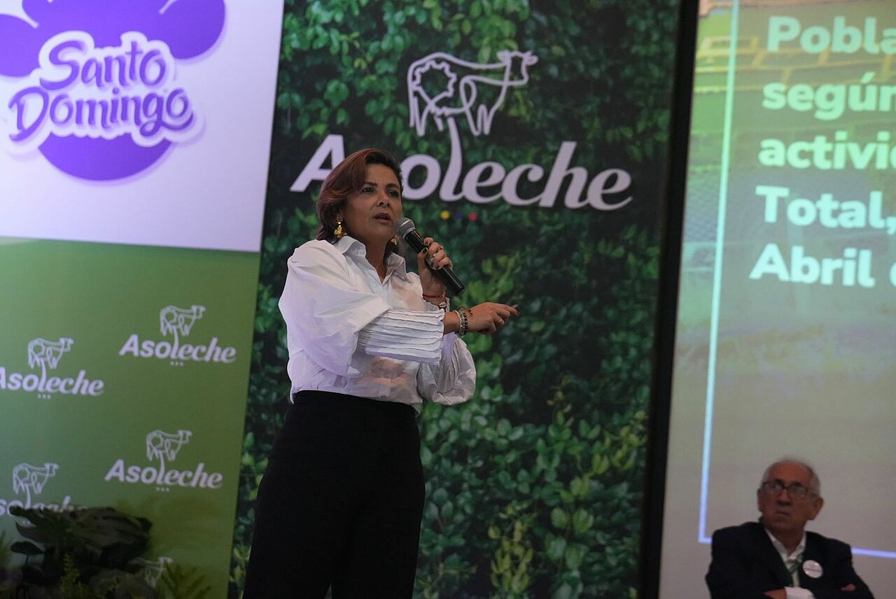 En la imagen aparece una mujer de pie en un escenario, sosteniendo un micrófono y hablando. Parece estar dando una presentación o un discurso. El escenario sugiere que podría tratarse de un evento corporativo, una conferencia o una reunión profesional similar.