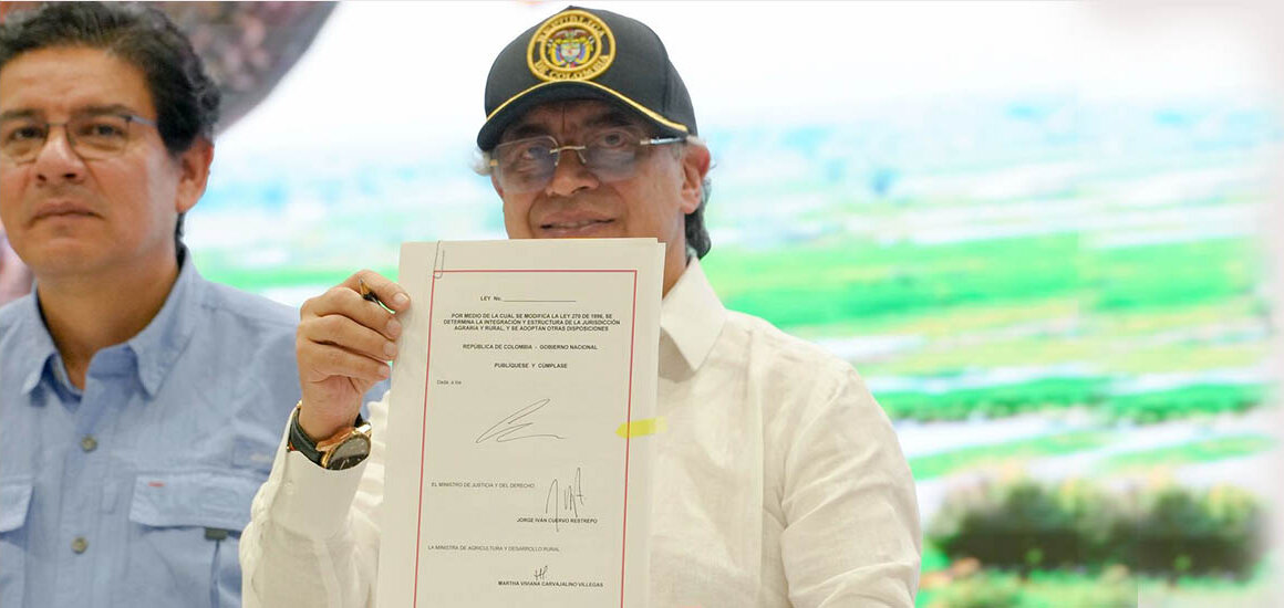 Dos personas están de pie en un espacio interior. La persona a la derecha viste camisa blanca, gafas y una gorra negra con un emblema oficial. Sostiene un documento frente a la cámara, mostrando claramente el texto y las firmas.