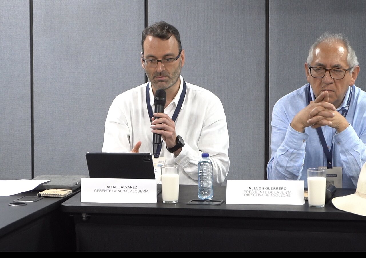 La imagen muestra a tres hombres sentados a una mesa, inmersos en una discusión. Uno de los hombres lleva gafas y parece estar hablando por un micrófono, posiblemente dirigiéndose a un público o participando en una mesa redonda. Los otros dos hombres también llevan gafas y parecen escuchar atentamente al orador.