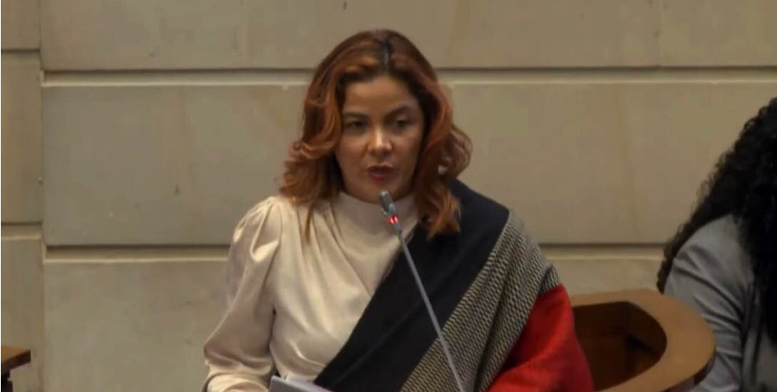 La imagen muestra a una mujer delante de un micrófono, dando un discurso o una presentación. Lleva una camisa blanca y un pañuelo negro, lo que añade un toque de elegancia a su aspecto. La mujer parece ser el centro de atención de la escena, ya que se dirige con confianza a su público.