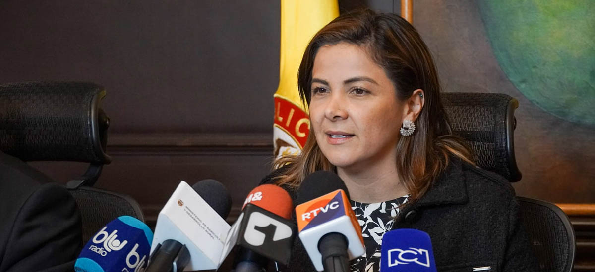 En la imagen se puede observar a la Ministra Jhenifer Mojica, dando una conferencia de prensa, con la bandera de Colombia de fondo