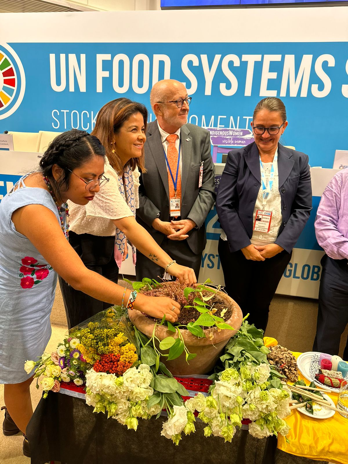 La imagen muestra a un grupo de personas reunidas en torno a una mesa, adornada con un gran cuenco lleno de diversas frutas y verduras. Los comensales discuten o hacen una presentación, y una de las mujeres deposita una hoja en el cuenco. La mesa también está decorada con un jarrón, lo que aumenta el atractivo visual de la escena.