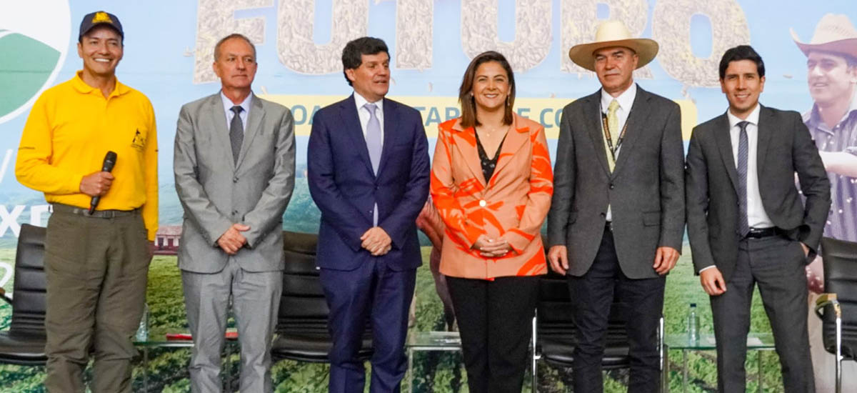 La imagen muestra a un grupo de cinco personas de pie unas junto a otras, todas con traje y corbata. El grupo está formado por tres hombres y dos mujeres, todos con atuendo profesional.  Al fondo se ven dos sillas, una a la izquierda y otra a la derecha del grupo. Las sillas parecen formar parte del entorno en el que está reunido el grupo.