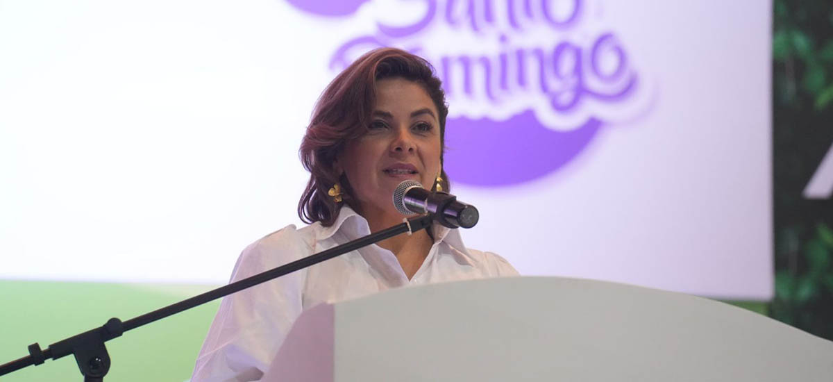 En la imagen se ve a la Ministra Jhenifer Mojica, de pie ante un podio con un micrófono, presumiblemente dando un discurso o una presentación. Lleva una blusa blanca y el pelo peinado de manera que le cae sobre los hombros.