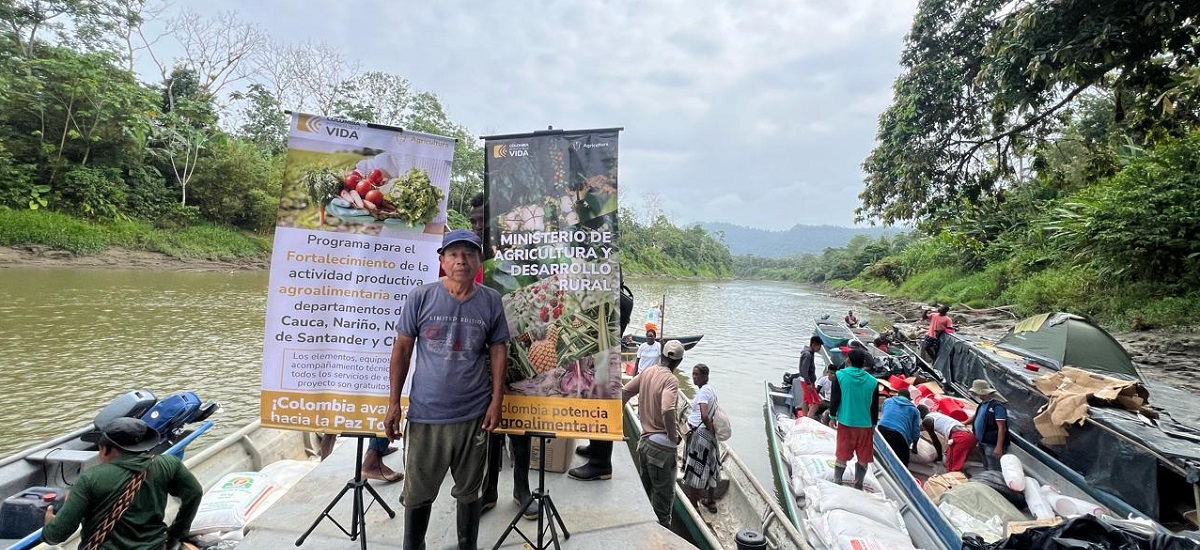 En la imagen se ve a una persona de pie en un barco con un río y una exuberante vegetación como telón de fondo. El cartel parece estar relacionado con un programa o iniciativa, posiblemente relacionados con la agricultura o el desarrollo rural, como indica el texto del cartel. El entorno parece tranquilo y natural, con árboles y follaje a orillas del río. El tiempo parece nublado y las aguas tranquilas.