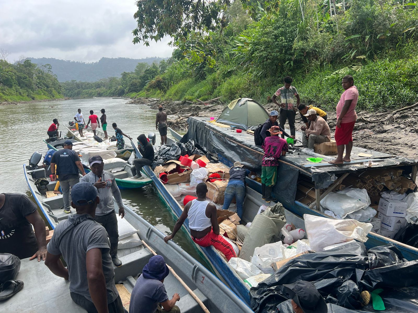 En la imagen se ve a un grupo de personas en la orilla de un río con varias barcas. Las embarcaciones parecen ser pequeñas, posiblemente motorizadas, y se están utilizando para el transporte o posiblemente para pescar.