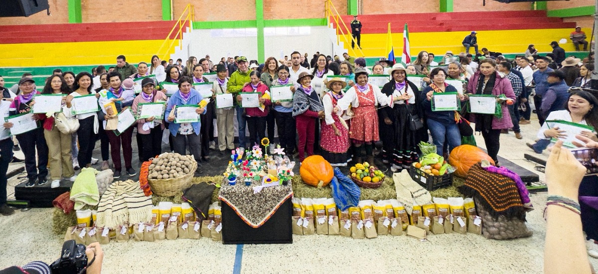 Esta imagen muestra a un grupo de personas reunidas en lo que parece ser un pabellón deportivo cubierto o un lugar grande similar. El grupo es diverso, con individuos de diversas edades y atuendos, lo que sugiere que se trata de un acto o reunión de la comunidad. Algunas personas sostienen certificados o premios, lo que indica que podrían haber recibido reconocimiento o premios por algún logro.
