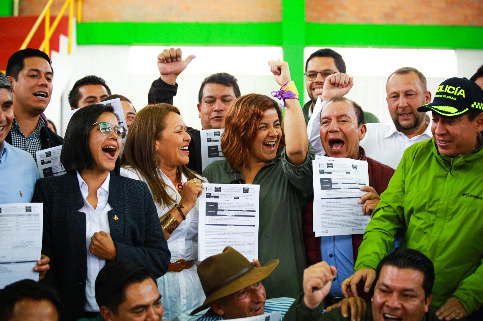 En la imagen, hay un grupo de personas que parecen estar celebrando o expresando una sensación de logro. Sostienen papeles que podrían ser documentos o certificados, lo que sugiere que podrían estar recibiendo premios, diplomas o reconocimiento por un logro. Las personas sonríen y levantan el puño, un gesto que suele asociarse con el triunfo o la solidaridad. El escenario parece un evento en un lugar cerrado, posiblemente una ceremonia o una reunión de algún tipo. El ambiente parece alegre y positivo.