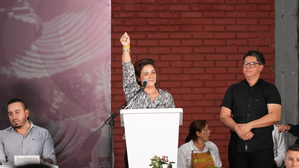En la imagen aparece una mujer de pie en un podio, levantando la mano derecha en un gesto que podría interpretarse como un saludo o una señal de agradecimiento. Parece estar hablando o dirigiéndose a un público, como sugiere la presencia de un micrófono en el podio.