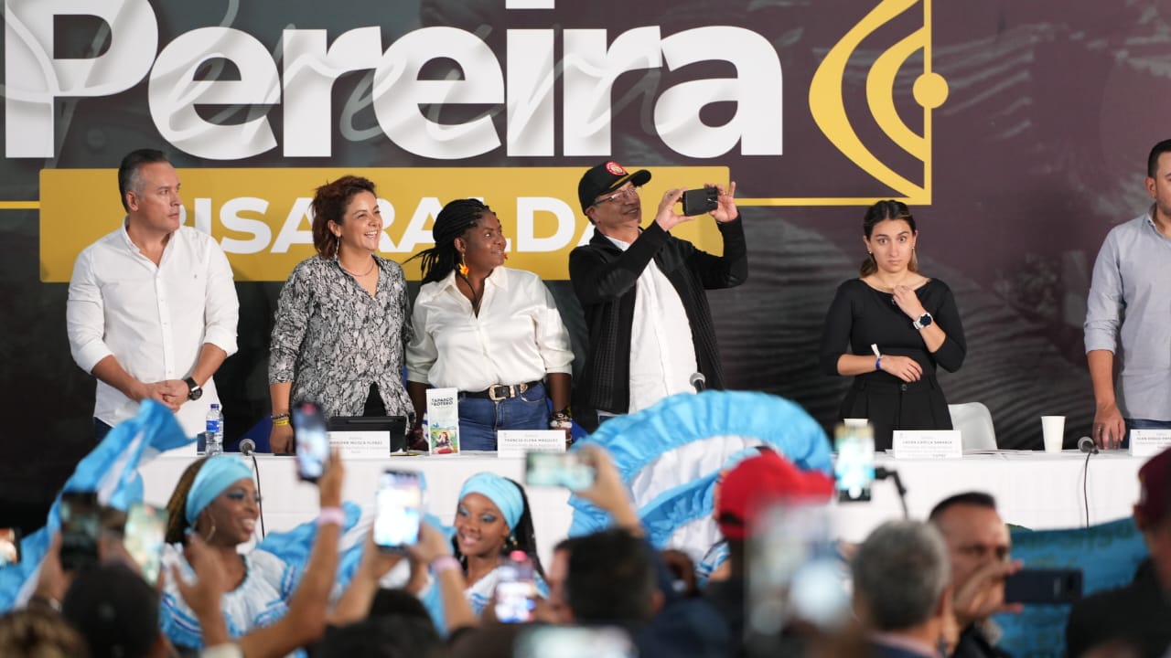 Esta imagen muestra a un grupo de personas de pie detrás de una mesa, probablemente en un acto público o una rueda de prensa. Parece que están posando para una foto, y algunas personas sostienen sus teléfonos para hacer fotos o grabar el momento.