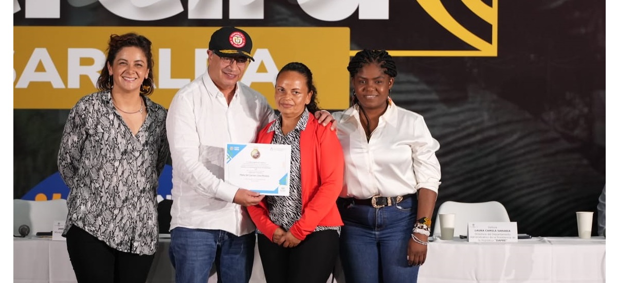 En la imagen hay cuatro personas juntas, probablemente en un acto o ceremonia. La persona del centro sostiene un certificado o un premio, lo que sugiere que se le está reconociendo un logro o una hazaña.