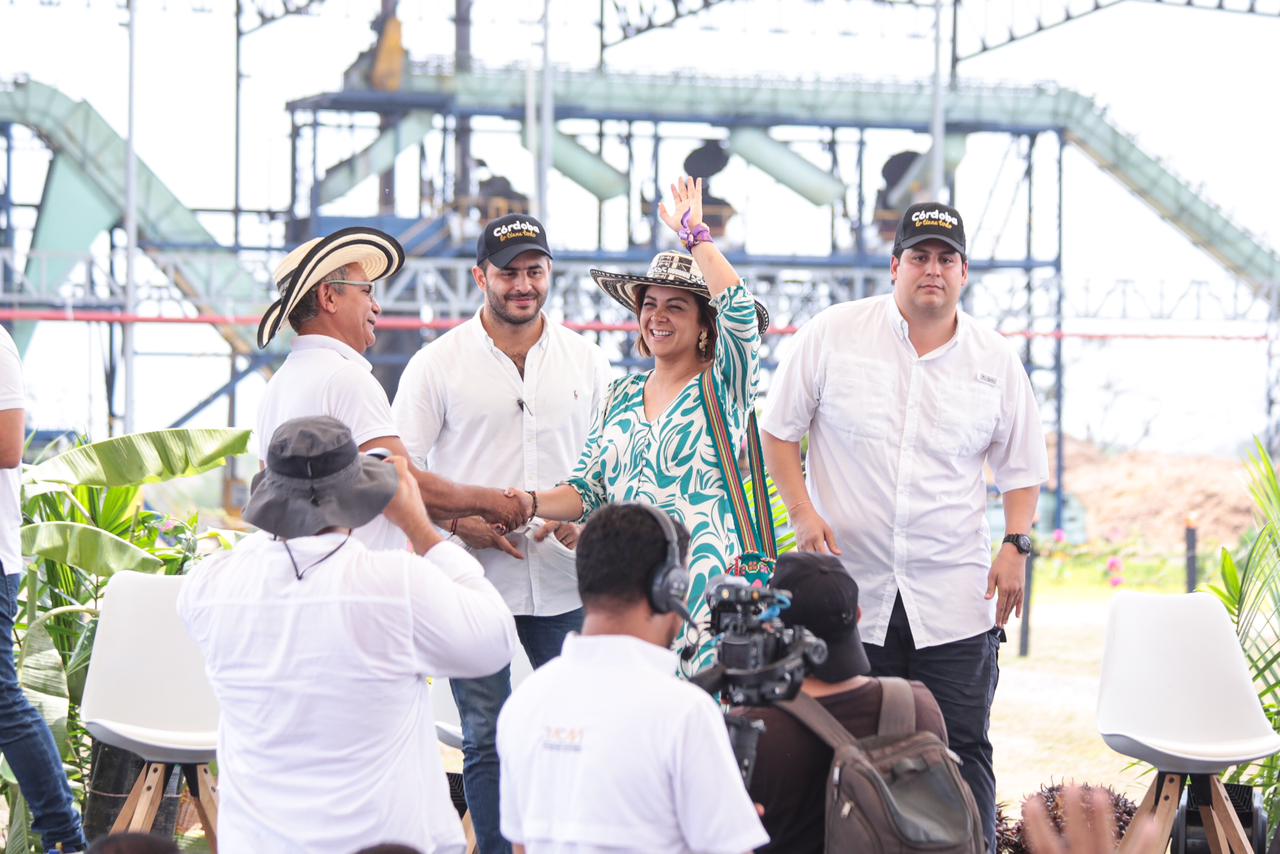 En la imagen, hay un grupo de personas reunidas al aire libre,  en un lugar con algún trabajo en curso. La figura central es la Ministra Jhenifer Mojica que sonríe y saluda con un sombrero y un top estampado. Está estrechando la mano de un hombre que lleva camisa blanca y sombrero.