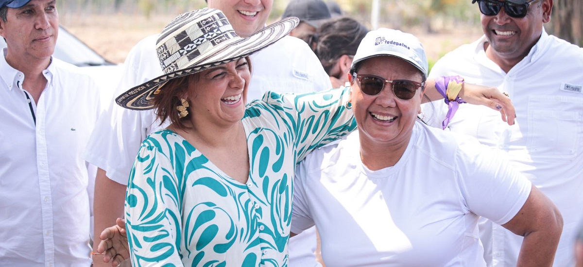 En la imagen hay dos mujeres que parecen estar alegres, sonriendo y riendo. La mujer de la izquierda es la ministra Jhenifer Mojica,  lleva un sombrero con un dibujo que parece un sol o una flor, y un top de colores con un dibujo similar. La mujer de la derecha lleva una gorra y un top blancos, y una banda morada alrededor de la muñeca. Parece que se están divirtiendo, posiblemente en un evento o reunión al aire libre.