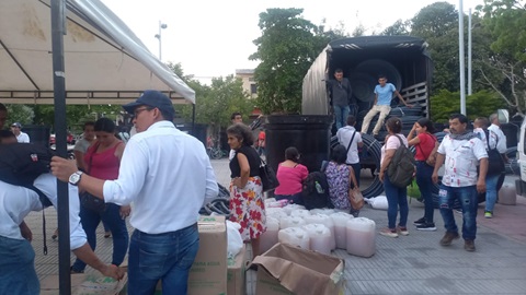 En la imagen, hay un grupo de personas reunidas en torno a un gran camión con la parte trasera abierta. El camión parece estar cargado con diversos artículos, posiblemente para su distribución o entrega.