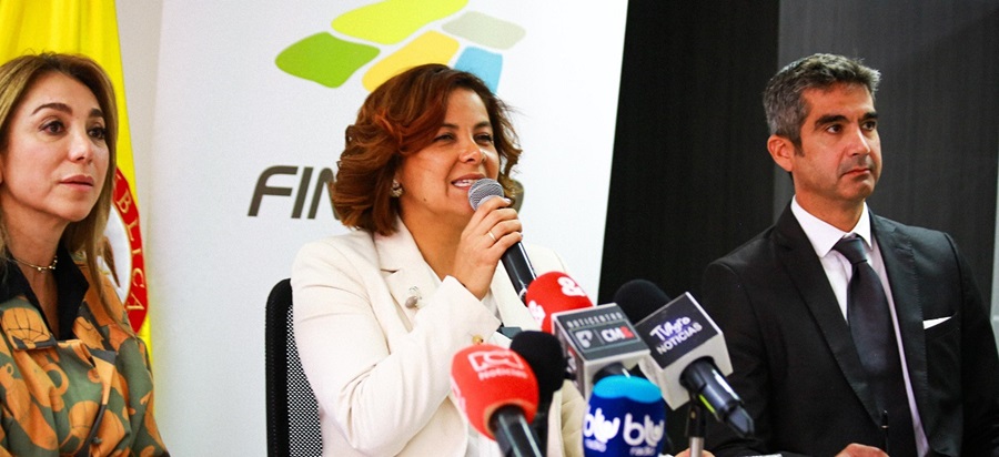 En la imagen se ve a la ministra Jhenifer Mojica  que parece estar hablando en una rueda de prensa o en un acto con los medios de comunicación. Sostiene un micrófono y parece dirigirse a los medios de comunicación, como indica la presencia de varios micrófonos frente a ella. Hay otras dos personas en el encuadre; una está sentada a su derecha, mirándola atentamente, y la otra está sentada a su izquierda, también mirándola.