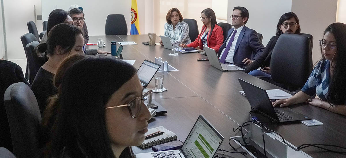 En la imagen se puede ver a varias personas reunidos en una sala de conferencias, todos con computadores portatiles y documentos, también se puede observar a la ministra Jhenifer Mojica escuchando atentamente.