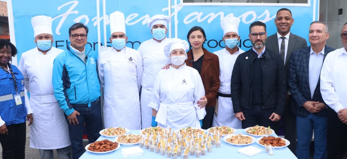 La imagen muestra un evento de carácter profesional o educativo en el ámbito culinario. En primer plano se ve una mesa con varios platos y recipientes pequeños que contienen aperitivos o muestras de comida cuidadosamente presentadas. Detrás de la mesa, un grupo de personas posa para la foto: algunos llevan uniforme de chef con gorro y mascarilla, lo que indica que son profesionales de cocina, mientras que otros están vestidos con ropa formal o casual, sugiriendo que son organizadores, invitados o representantes institucionales.