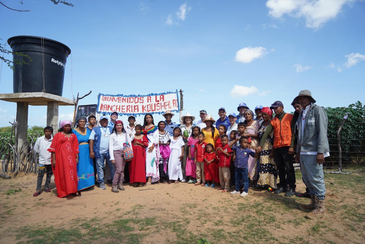 La imagen muestra a un grupo numeroso de personas de pie en un campo, posando para una foto. Hay al menos 14 personas en el grupo, algunas de ellas con sombrero. Están de pie delante de un cartel, que probablemente sea el punto central de la reunión. Las personas están repartidas por el campo, algunas más cerca de la pancarta y otras más lejos.