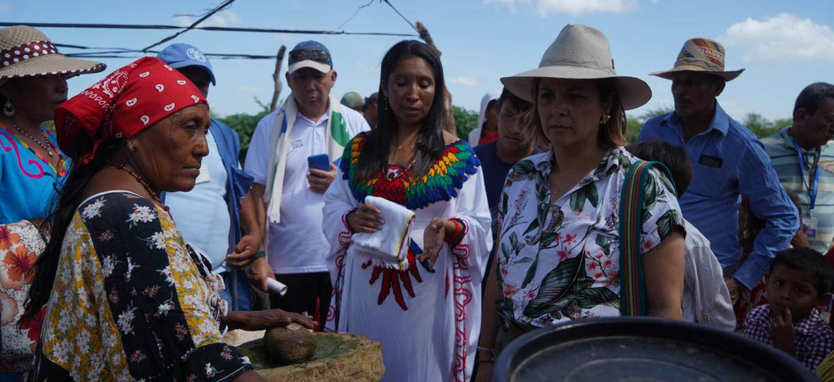 n la escena hay varias personas, algunas con sombrero y otras vestidas con ropas tradicionales. Una mujer está de pie frente a un hombre que sostiene un teléfono móvil. El grupo parece estar conversando u observando algo juntos.