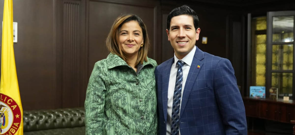 La imagen muestra a un hombre y una mujer de pie, uno junto al otro, posando para una foto. El hombre lleva traje y corbata, mientras que la mujer viste una chaqueta verde. Ambos sonríen y parecen disfrutar del momento.