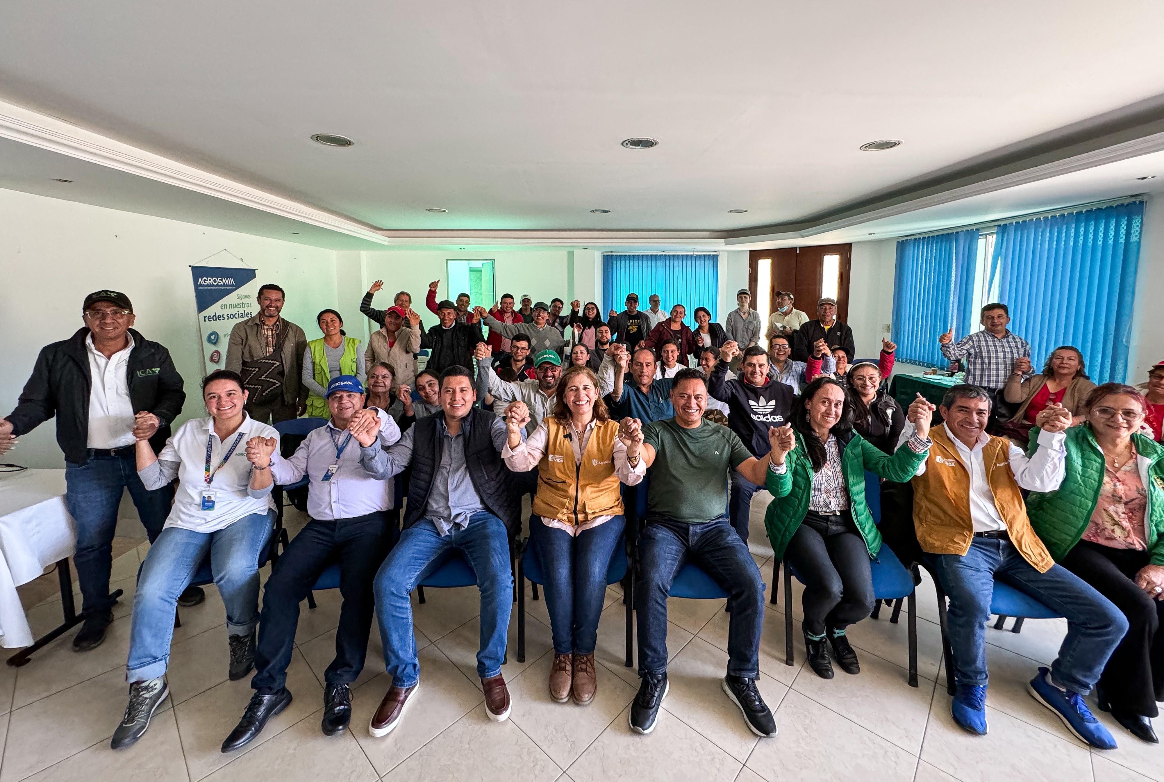 En la imagen, hay un grupo de personas sentadas en fila, con las manos levantadas en un gesto que sugiere que están celebrando o animando.  Detrás de ellos, hay un grupo más grande de personas de pie que también levantan las manos, lo que refuerza el ambiente de celebración. El escenario parece ser un espacio interior para eventos, posiblemente una sala de conferencias o un salón utilizado para reuniones o presentaciones.