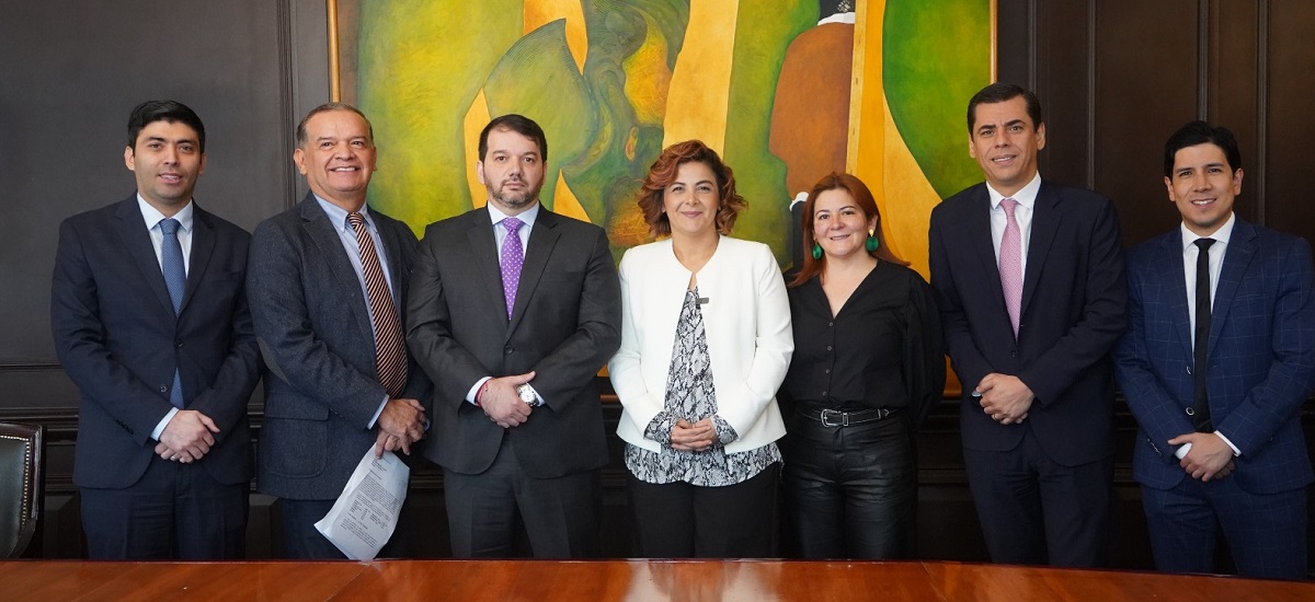 La imagen muestra a un grupo de siete personas, todas ellas vestidas con traje de negocios, de pie frente a un cuadro. Parecen posar para una foto de grupo, y algunos de ellos sonríen. El grupo está formado por hombres y mujeres, todos con corbata.