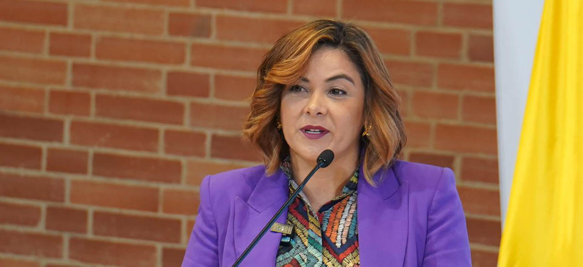 La imagen muestra a una mujer delante de un micrófono, con una chaqueta morada y una camisa de colores. Parece estar dando un discurso o una presentación, ya que está hablando por el micrófono. La mujer es el foco principal de la escena, y su atuendo y postura sugieren que está segura de sí misma y comprometida con su presentación.