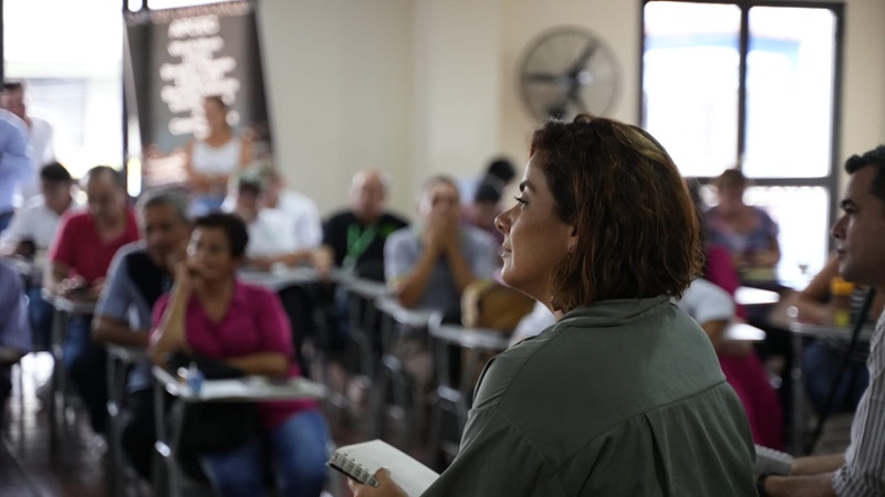 En la imagen se ve a la ministra Jhenifer Mojica de pie frente a un grupo de personas que parecen estar sentadas en un aula o una sala de conferencias. Sostiene lo que parece un libro o un cuaderno, lo que sugiere que podría estar tomando nota de comentarios importantes en la reunion.