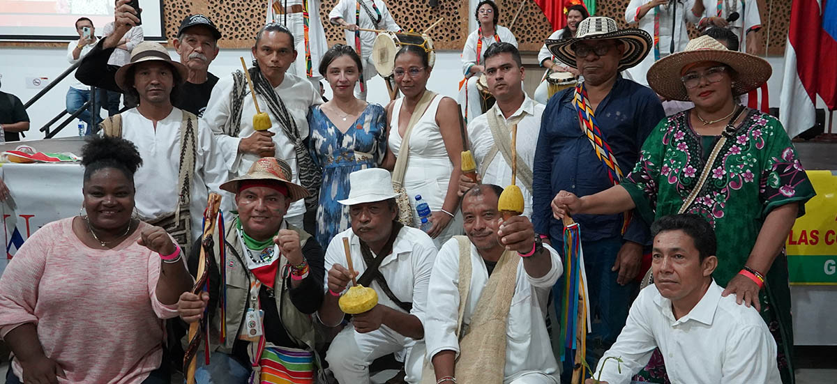 La imagen muestra a un grupo de personas reunidas en un espacio interior, probablemente en el marco de un evento cultural o comunitario. Se observa una diversidad de vestimenta: algunos llevan atuendos tradicionales con colores vivos, sombreros y accesorios que evocan raíces indígenas o regionales, mientras otros visten ropa más contemporánea. Varias personas sostienen objetos ceremoniales como bastones o recipientes, lo que sugiere un componente ritual o simbólico dentro de la reunión.