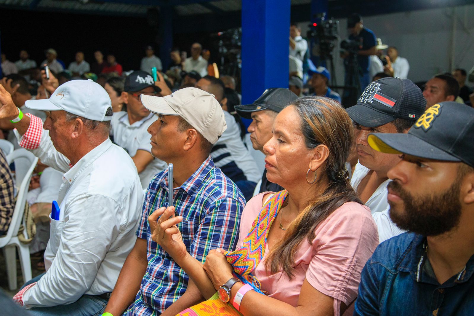 En la imagen se ve a un grupo de personas sentadas en lo que parece ser un lugar cerrado, posiblemente una conferencia o un evento. Miran atentamente hacia el frente, donde podría haber un orador o una presentación. Los asistentes van vestidos con ropa informal y algunos llevan sombreros, lo que sugiere un ambiente relajado o informal